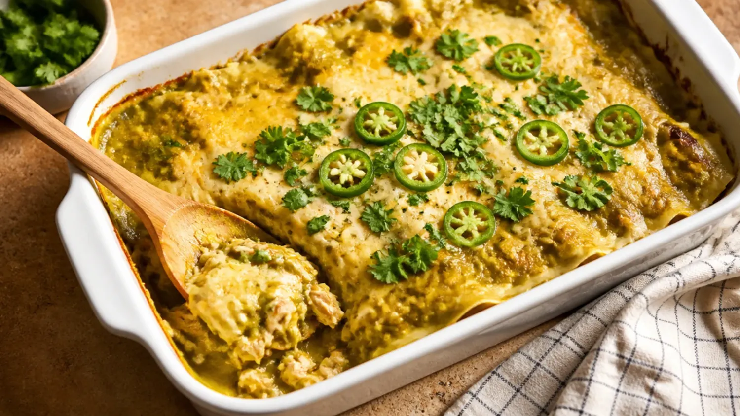 Overhead shot of a bubbling green chicken enchilada casserole in a white baking dish, topped with melted cheese, sour cream drizzle, chopped coriander and sliced jalapeños