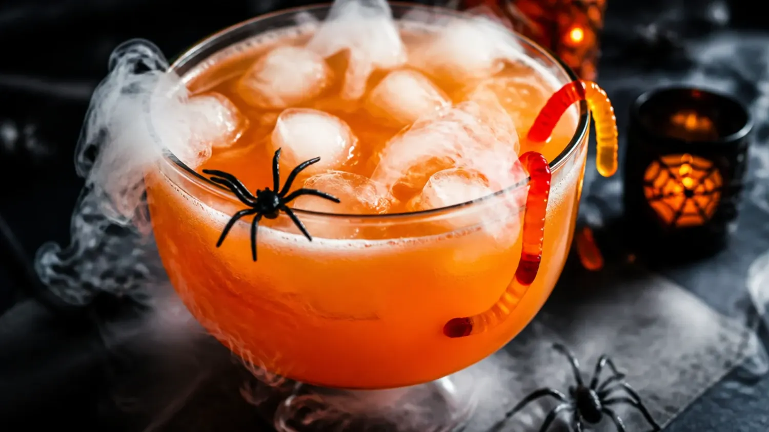 A glowing green Halloween witches' brew punch in a black cauldron bowl, surrounded by candy eyeballs, gummy worms and dry ice fog on a dark table.