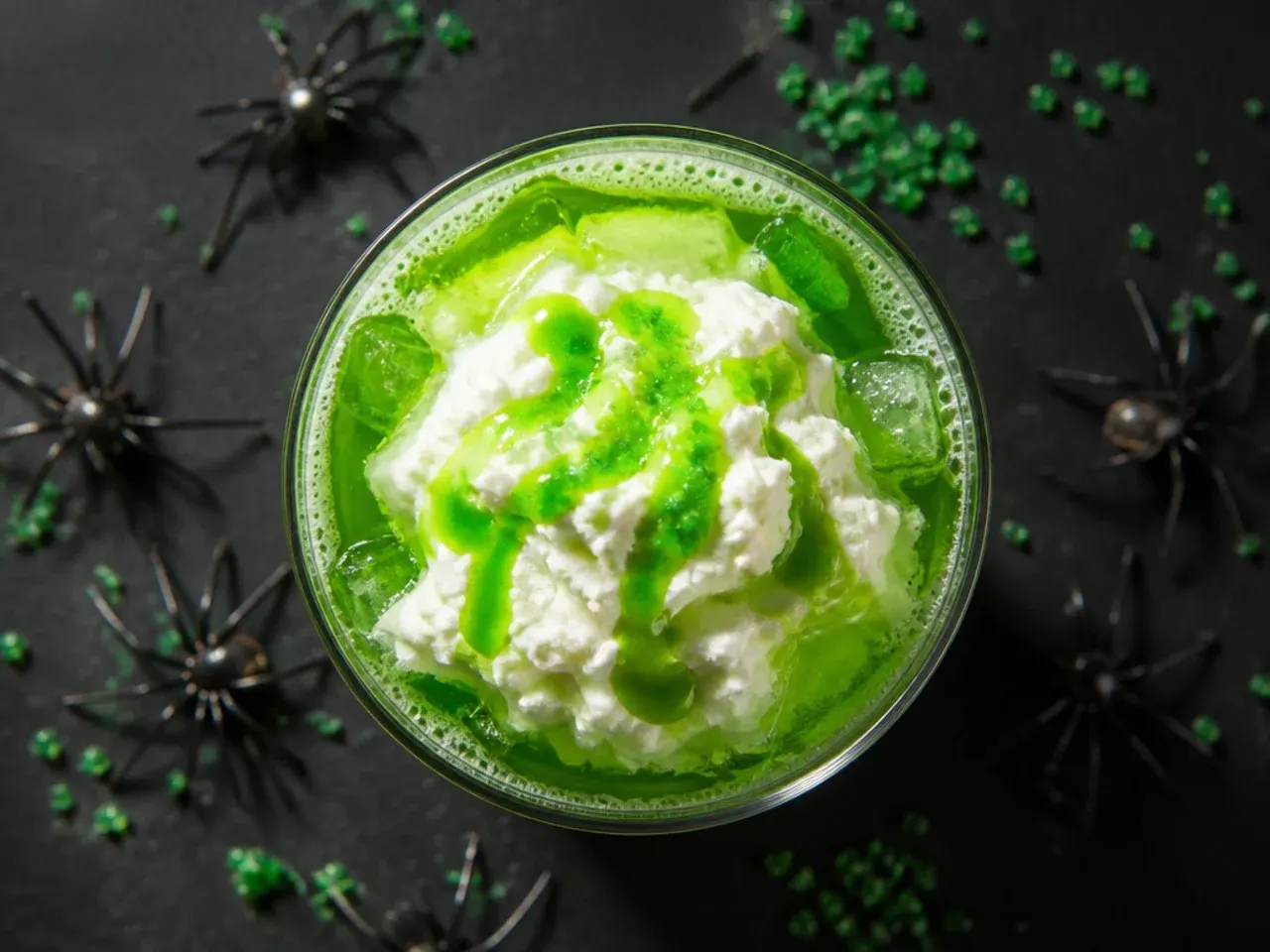 Tall glass of green iced witches brew drink with a plastic spider ring on the rim, surrounded by ice.
