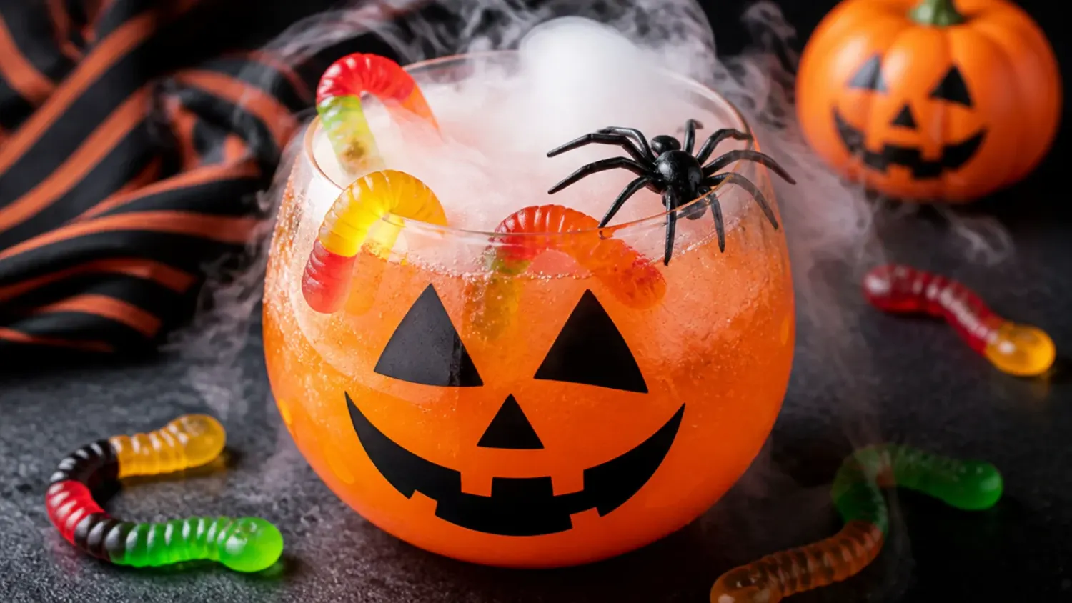 A table spread of colourful halloween drinks for kids in glass cups with gummy worms, candy eyeballs and orange straws