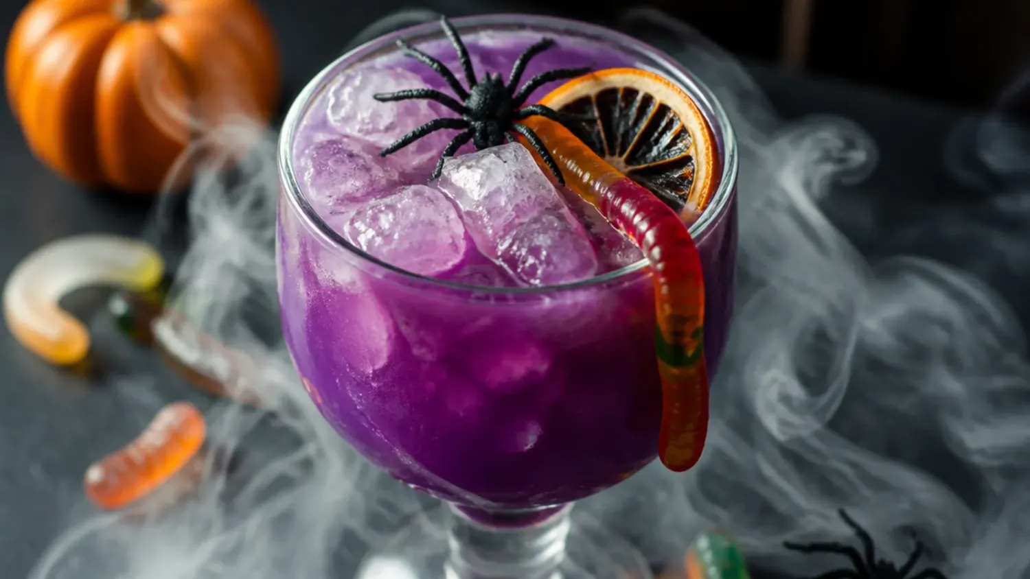 A glowing orange and purple Halloween mixed drink in a stemmed glass, garnished with a lime wheel and black sugar rim, surrounded by mini pumpkins and dry ice fog